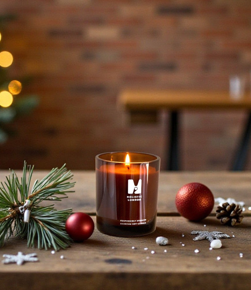 Candle in a brown glass jar with a brand logo, surrounded by Christmas decorations on a wooden table.